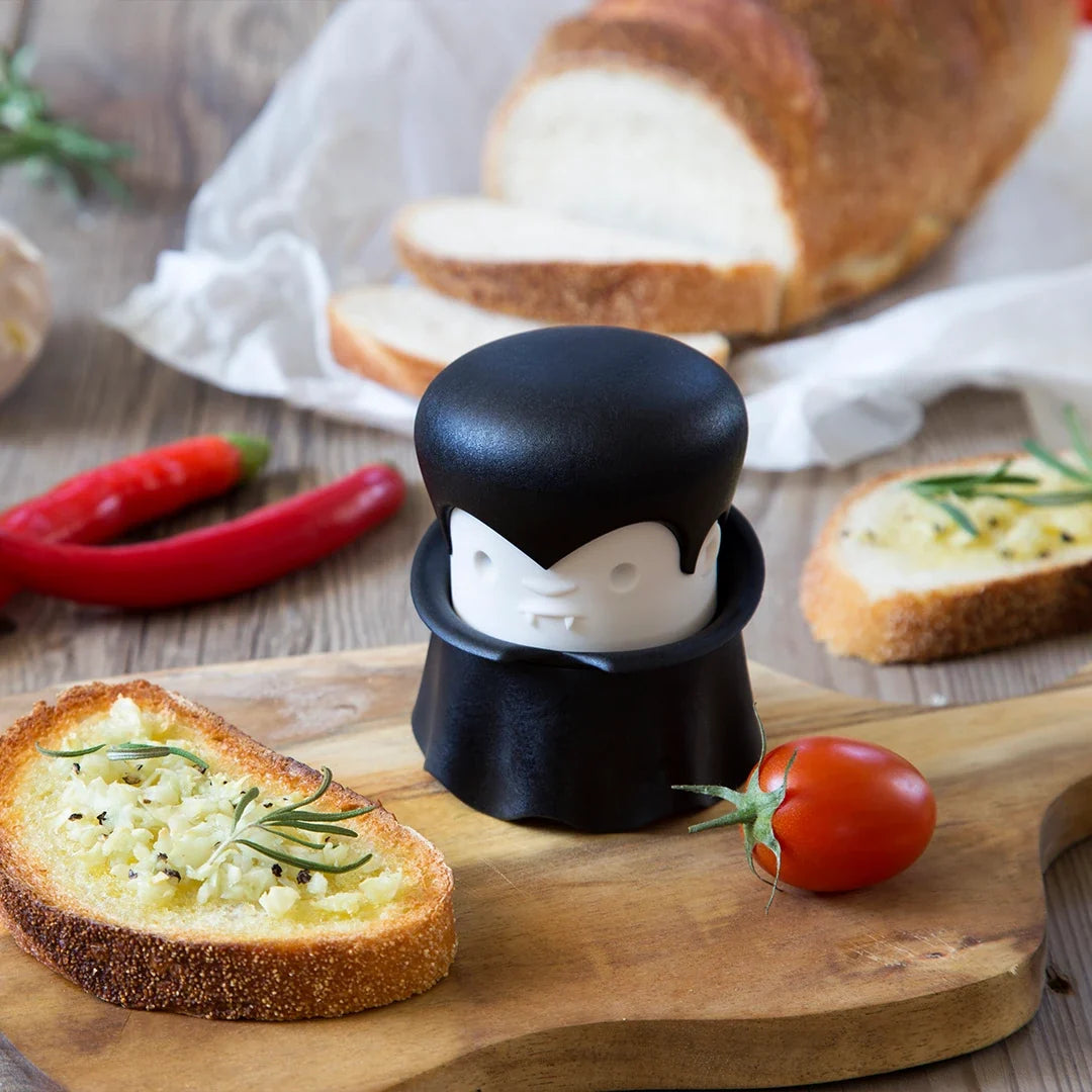 The Gracula garlic crusher sits by toasted bread, cheese, rosemary, and cherry tomatoes on a wooden table.